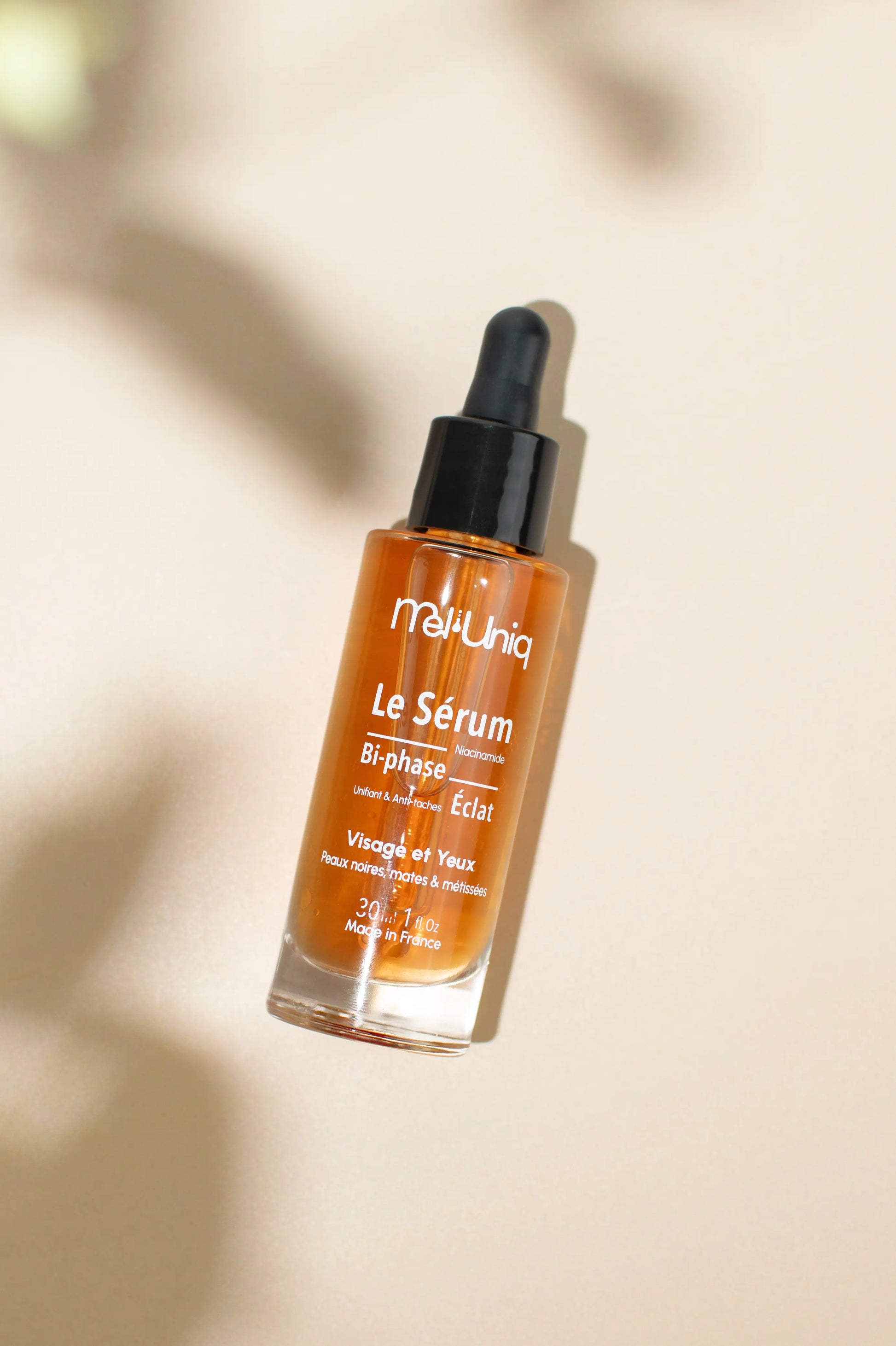 Flacon du sérum bi-phase éclat Mel’Uniq sur fond beige avec jeu d’ombres naturelles