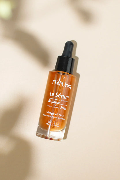 Flacon du sérum bi-phase éclat Mel’Uniq sur fond beige avec jeu d’ombres naturelles
